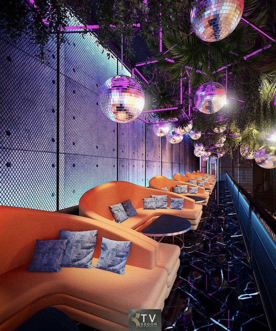 Khu lounge được thiết kế theo phong cách hiện đại – thư giãn, nổi bật với dãy sofa bo cong màu cam ấm, tạo cảm giác thoải mái và sang trọng. Không gian được nâng tầm nhờ hệ trần trang trí cây xanh kết hợp đèn cầu gương phản chiếu ánh sáng, mang lại hiệu ứng thị giác sinh động về đêm. Ánh sáng LED tím – xanh được bố trí tinh tế, giúp khu vực lounge vừa riêng tư vừa cuốn hút, phù hợp cho nhóm khách trò chuyện, thưởng thức đồ uống và thư giãn sau giờ làm việc. 