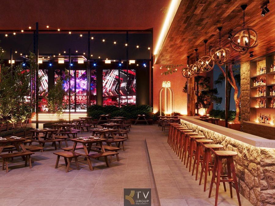 Không gian Beer Garden Phú Quốc – Kiên Giang được thiết kế theo phong cách mở, kết hợp hài hòa giữa vật liệu tự nhiên và ánh sáng nghệ thuật. Khu vực quầy bar nổi bật với đá ốp thô mộc, kệ trưng bày bia – rượu sang trọng, ghế bar gỗ cao tạo điểm tụ họp sôi động. Trần ốp gỗ cùng hệ đèn thả trang trí mang lại cảm giác ấm cúng nhưng vẫn hiện đại. Bên ngoài là khu bàn dài bố trí linh hoạt, phù hợp cho nhóm đông, tiệc tối và sự kiện âm nhạc 